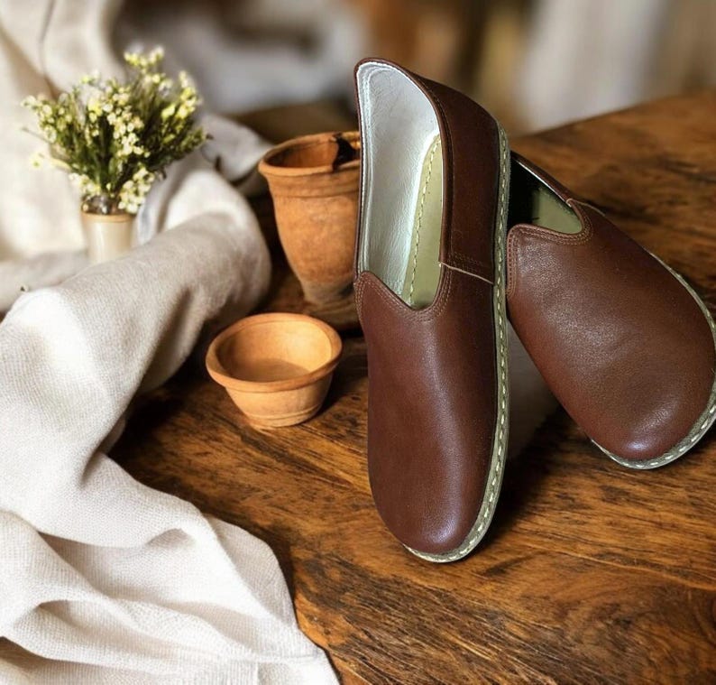 May include: A pair of brown leather slip-on shoes with white stitching around the edges. The shoes are on a wooden surface.