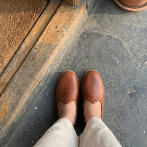 May include: A pair of smooth, cognac-colored leather slip-on shoes with a rounded toe and a low-profile design, worn with light beige linen trousers. The shoes feature a simple, clean construction with visible stitching along the sole edge.