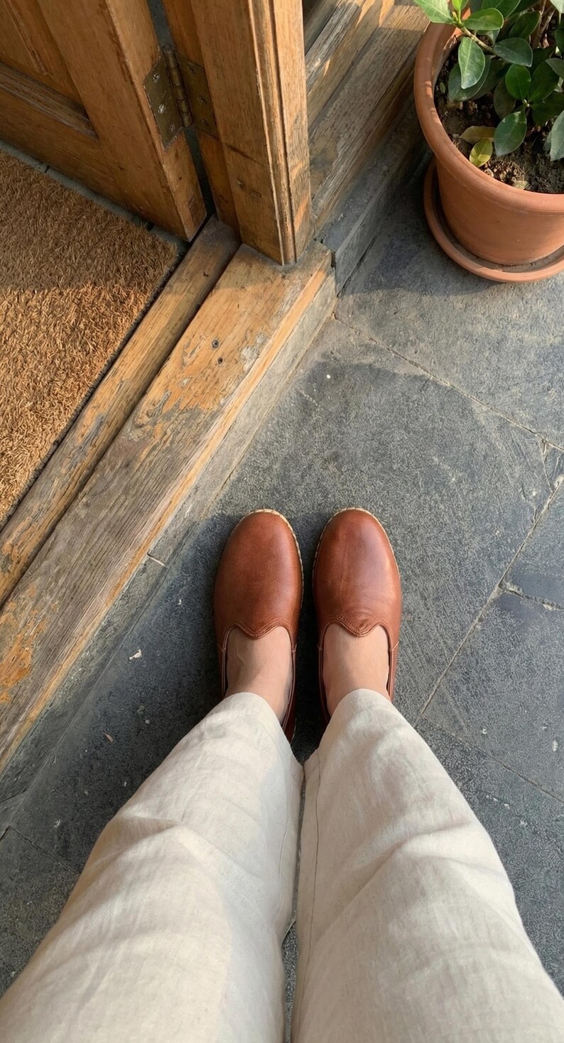 May include: A pair of smooth, cognac-colored leather slip-on shoes with a rounded toe and a low-profile design, worn with light beige linen trousers. The shoes feature a simple, clean construction with visible stitching along the sole edge.