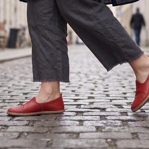 Puede incluir: Zapatos rojos de cuero sin cordones con suela color canela. Los zapatos se usan con pantalones grises con dobladillo deshilachado. La imagen est&aacute; tomada en una calle adoquinada.