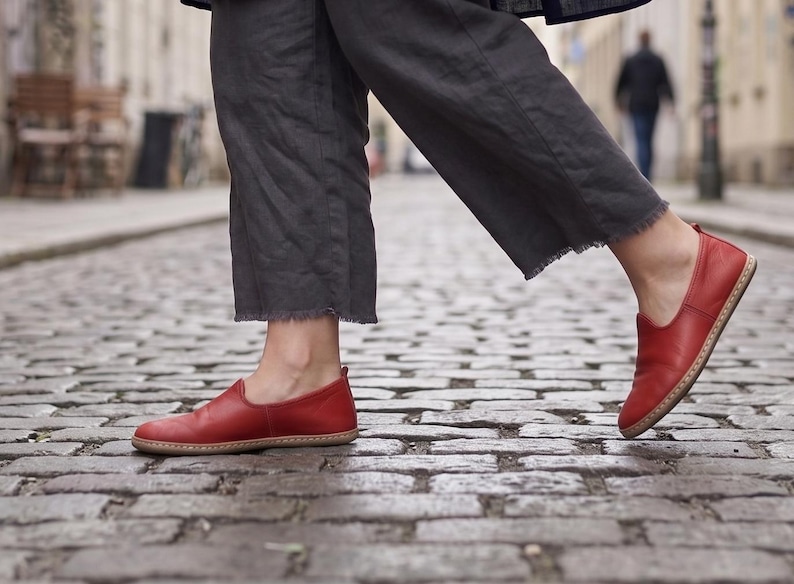 Puede incluir: Zapatos rojos de cuero sin cordones con suela color canela. Los zapatos se usan con pantalones grises con dobladillo deshilachado. La imagen est&aacute; tomada en una calle adoquinada.