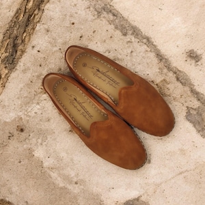 K&ouml;nnte beinhalten: Ein Paar braune Wildleder-Loafer mit runder Spitze und Ziern&auml;hten. Die Schuhe zum Hineinschl&uuml;pfen haben eine Innensohle aus Leder. Der Markenname ist auf der Innensohle aufgedruckt. Die Schuhe werden auf einer strukturierten, hellen Oberfl&auml;che pr&auml;sentiert.