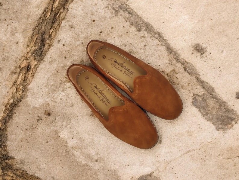 K&ouml;nnte beinhalten: Ein Paar braune Wildleder-Loafer mit runder Spitze und Ziern&auml;hten. Die Schuhe zum Hineinschl&uuml;pfen haben eine Innensohle aus Leder. Der Markenname ist auf der Innensohle aufgedruckt. Die Schuhe werden auf einer strukturierten, hellen Oberfl&auml;che pr&auml;sentiert.