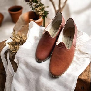 May include: A pair of slip-on shoes in a warm, reddish-brown tone with a rounded toe and a low-cut opening. The interior soles feature a printed logo that reads "Charwood Handmade Footwear" with a size indicator. The edges are stitched with light-colored thread.