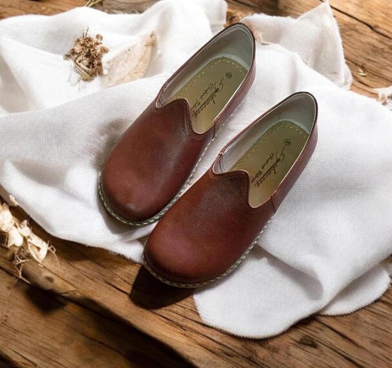 May include: A pair of brown leather slip-on shoes with a pointed toe and a simple design. The shoes have a white leather lining and a white leather sole. The brand name "Alessandro" is printed on the inside of the shoes.