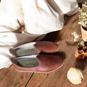 May include: A pair of brown leather slip-on shoes with a stitched sole, resting on a wooden surface. The shoes have a simple, classic design. The interior of the shoe has the text "sharvey Handmade Footwear".