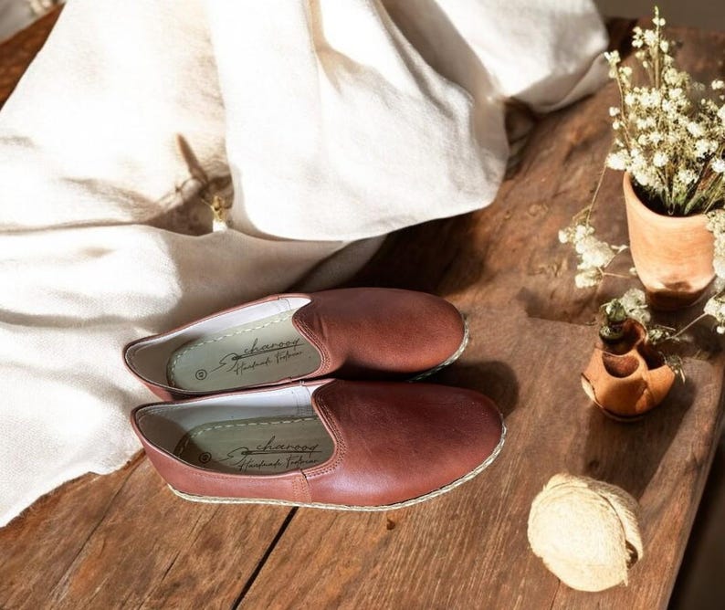 May include: A pair of brown leather slip-on shoes with a stitched sole, resting on a wooden surface. The shoes have a simple, classic design. The interior of the shoe has the text "sharvey Handmade Footwear".