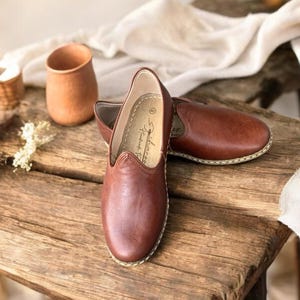 May include: A pair of brown slip-on shoes with a rounded toe and visible light-colored stitching along the sole. The interior of the shoe features a printed logo and the number 43. The shoes have a smooth finish and a low-profile design.