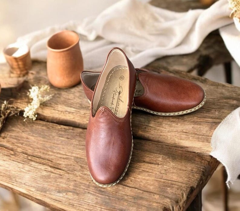 May include: A pair of brown slip-on shoes with a rounded toe and visible light-colored stitching along the sole. The interior of the shoe features a printed logo and the number 43. The shoes have a smooth finish and a low-profile design.