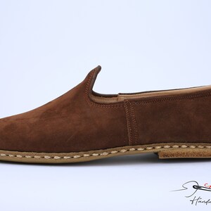 May include: A brown suede slip-on shoe with a rounded toe and a low-profile silhouette. The shoe features a tan-colored sole with visible light-colored stitching along the edge. The logo for Charooq Handmade Footwear is visible in the corner.