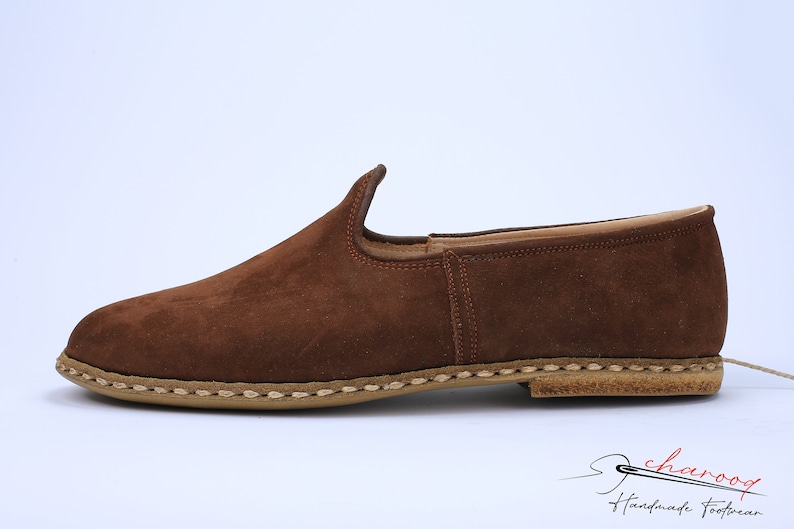 May include: A brown suede slip-on shoe with a rounded toe and a low-profile silhouette. The shoe features a tan-colored sole with visible light-colored stitching along the edge. The logo for Charooq Handmade Footwear is visible in the corner.