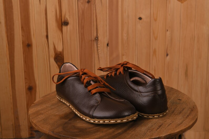 May include: A pair of brown leather shoes with orange laces. The shoes have a stitched seam around the sole and are sitting on a wooden surface.
