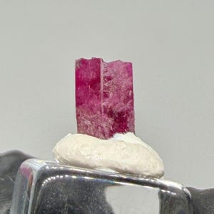 May include: A ring featuring a clear, square-shaped glass or crystal setting. A small, vibrant magenta-pink raw mineral crystal is mounted on top of the clear square using a white, textured adhesive base. The ring band is not visible.