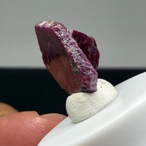 Op de afbeelding: Een ruw, diep magentakleurig kristalexemplaar met een platte, plaatachtige structuur en een ruw, getextureerd oppervlak. Het kristal vertoont natuurlijke facetten en onregelmatige randen, met een rijke, verzadigde rood-paarse tint door de hele vorm.