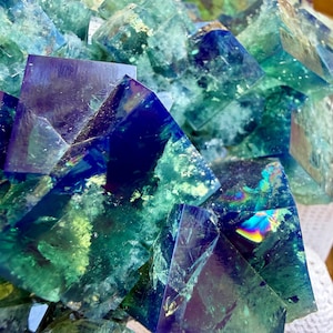 May include: A cluster of translucent fluorite crystals featuring sharp cubic formations in deep shades of royal blue, emerald green, and teal. The mineral specimen displays internal fractures, reflective surfaces, and vibrant rainbow light refractions.