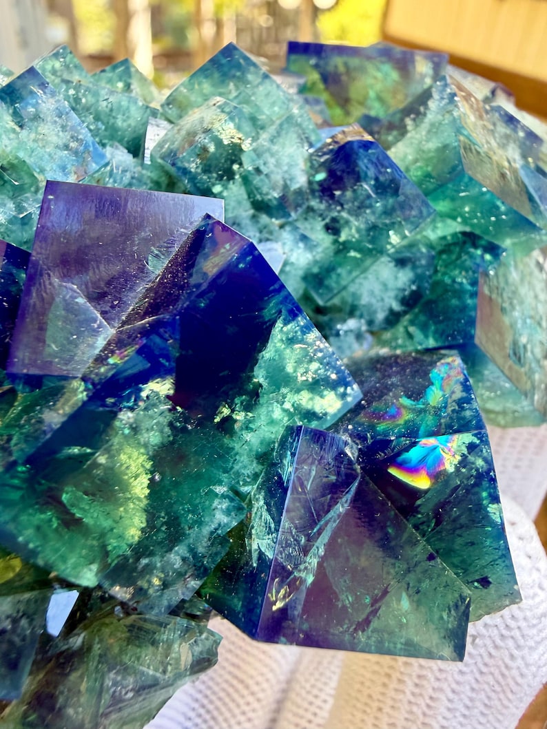 May include: A cluster of translucent fluorite crystals featuring sharp cubic formations in deep shades of royal blue, emerald green, and teal. The mineral specimen displays internal fractures, reflective surfaces, and vibrant rainbow light refractions.
