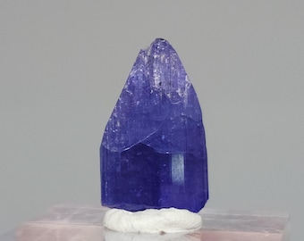 May include: A vibrant, faceted tanzanite crystal, showcasing a deep blue-violet hue. The gemstone is mounted on a white, fibrous base, set against a soft, neutral background. The crystal's surface reflects light, highlighting its natural structure.