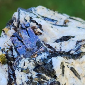 May include: Close-up of a mineral specimen with a mix of colors. The rock features a base of white and gray, with dark, almost black, inclusions. Patches of iridescent blue and purple crystals are visible, adding a striking contrast.