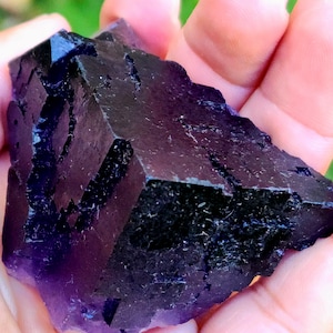 May include: A dark purple fluorite crystal with a complex, geometric structure. The crystal has a deep, almost black color in some areas, with lighter purple hues throughout. The surface has a rough, textured appearance, with visible crystalline formations.