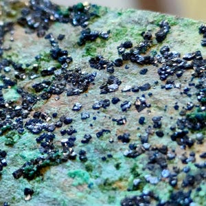 May include: Close-up of a mineral specimen with a rough, textured surface. The stone has a base color of tan and green, with clusters of dark, metallic-looking crystals scattered across it. The crystals have a reflective, almost iridescent quality.