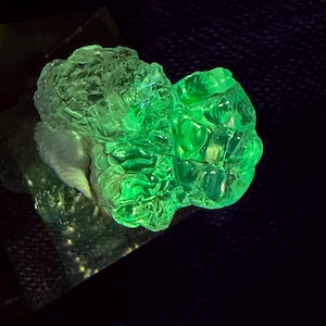 May include: A raw, translucent mineral specimen exhibiting a vibrant, glowing neon green fluorescence. The structure is bulbous and irregular with a glassy, crystalline texture, appearing to emit light against a dark, non-reflective background.