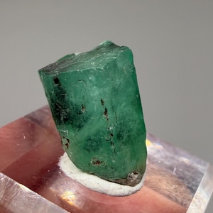 May include: A raw, hexagonal emerald crystal with a deep green hue and visible natural inclusions. The gemstone features a translucent, prismatic structure with vertical striations along its sides and a rough, unpolished surface texture.