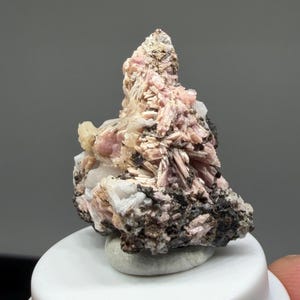Pode incluir: Um espécime mineral bruto apresentando um aglomerado de cristais rosa-pálidos em forma de agulha, misturados com estruturas cristalinas brancas translúcidas e rocha de matriz cinzenta escura e texturizada. A formação tem uma forma irregular.