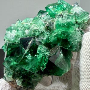 May include: A cluster of vibrant green fluorite crystals featuring sharp, cubic geometric formations with translucent surfaces and deep emerald hues. The mineral specimen displays a complex, multi-faceted structure with varying levels of clarity and light reflection across its jagged, crystalline edges.