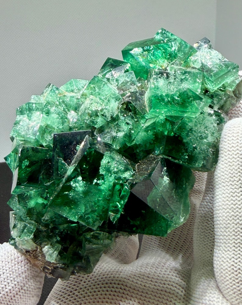 May include: A cluster of vibrant green fluorite crystals featuring sharp, cubic geometric formations with translucent surfaces and deep emerald hues. The mineral specimen displays a complex, multi-faceted structure with varying levels of clarity and light reflection across its jagged, crystalline edges.