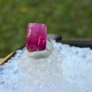 Op de afbeelding: Een levendige, rechthoekige, roze edelsteen, mogelijk toermalijn, wordt getoond op een kleine, lichtgekleurde, afgeronde basis. De edelsteen staat tegen een wazige groene en witte achtergrond, waarbij de kleur en vorm van de kristal worden benadrukt.