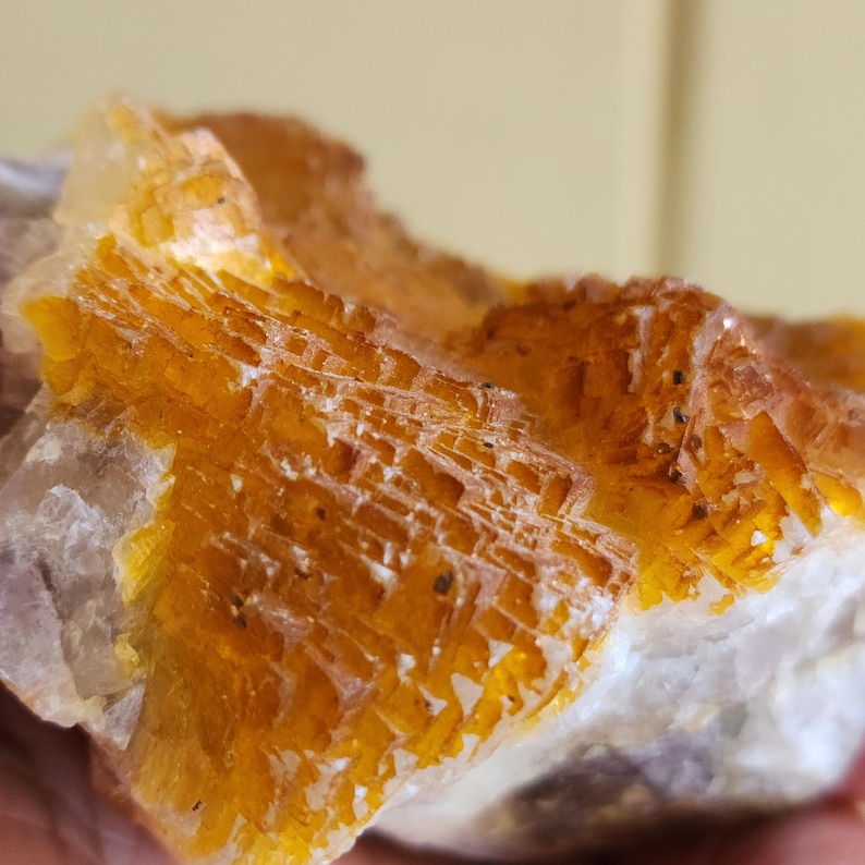 May include: Close-up of a rough, natural mineral specimen. The stone features a layered, crystalline structure with shades of orange, yellow, and white. The texture is rough and uneven, with visible lines and patterns throughout the surface.