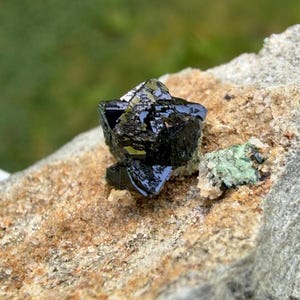 May include: Close-up of a dark, lustrous black mineral crystal formation. The crystal is sharply defined with multiple facets, set against a rough, tan and gray rock matrix. A small patch of green mineral is also present.