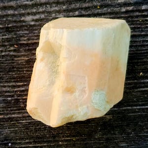 Puede incluir: Una gema en bruto sin pulir con forma rectangular. La piedra tiene un color melocotón pálido con áreas blancas y amarillas. La superficie tiene una textura rugosa y natural. La gema se encuentra sobre un fondo de vetas de madera oscura.