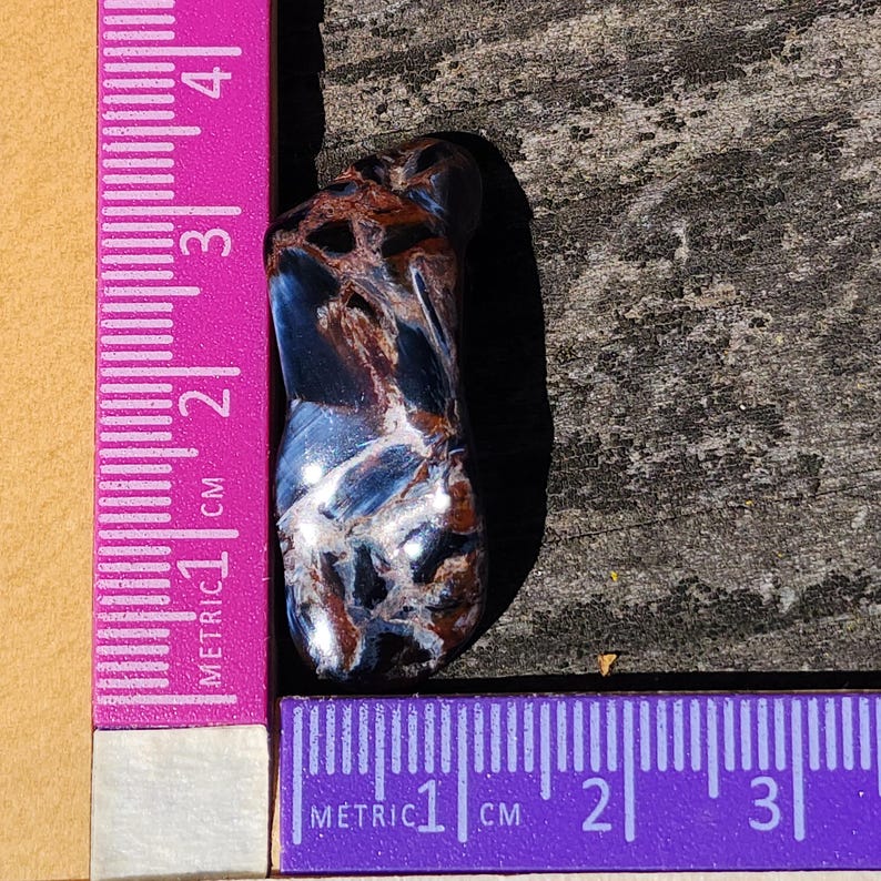 May include: A blue, brown, and white tiger's eye gemstone, measuring approximately 2 cm in length, is placed on a wooden surface next to a pink ruler.