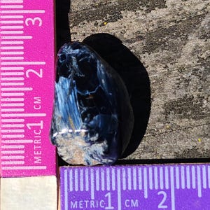 K&ouml;nnte beinhalten: Ein blauer und brauner Tigerauge-Edelstein mit einer rauen, nat&uuml;rlichen Oberfl&auml;che. Der Stein ist etwa 1 cm lang und wird an einem Lineal gemessen.