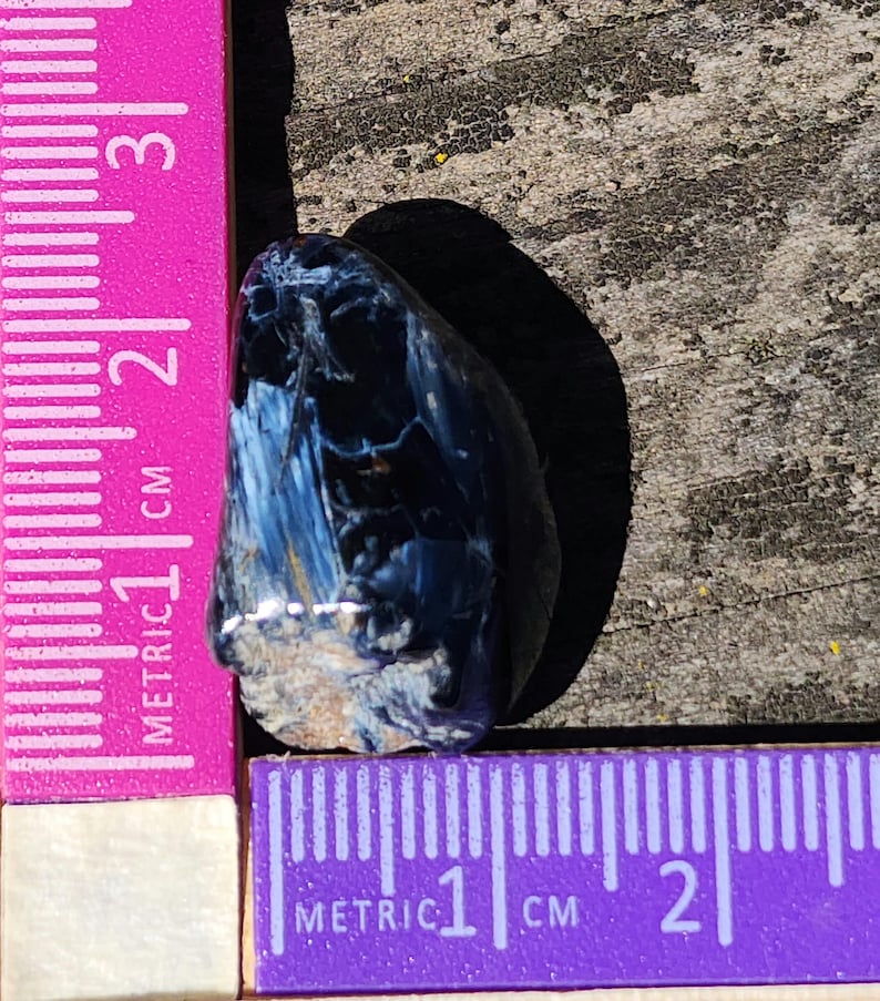 K&ouml;nnte beinhalten: Ein blauer und brauner Tigerauge-Edelstein mit einer rauen, nat&uuml;rlichen Oberfl&auml;che. Der Stein ist etwa 1 cm lang und wird an einem Lineal gemessen.
