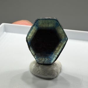 K&ouml;nnte beinhalten: Ein sechseckiger Edelstein mit blauen und gr&uuml;nen Farbt&ouml;nen, der auf einem kleinen, wei&szlig;en, strukturierten Sockel ruht. Der Edelstein hat ein dunkles Inneres und eine polierte Oberfl&auml;che. Das Bild zeigt nat&uuml;rliche Mineralien und Kristalle.