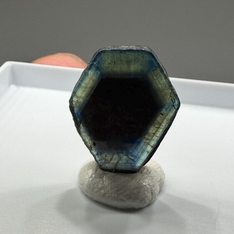 K&ouml;nnte beinhalten: Ein sechseckiger Edelstein mit blauen und gr&uuml;nen Farbt&ouml;nen, der auf einem kleinen, wei&szlig;en, strukturierten Sockel ruht. Der Edelstein hat ein dunkles Inneres und eine polierte Oberfl&auml;che. Das Bild zeigt nat&uuml;rliche Mineralien und Kristalle.