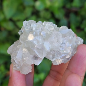 May include: A cluster of clear, crystalline minerals, likely a raw gemstone or mineral specimen. The crystals are translucent and have a slightly rough texture. The background is a blurred green, suggesting an outdoor setting.