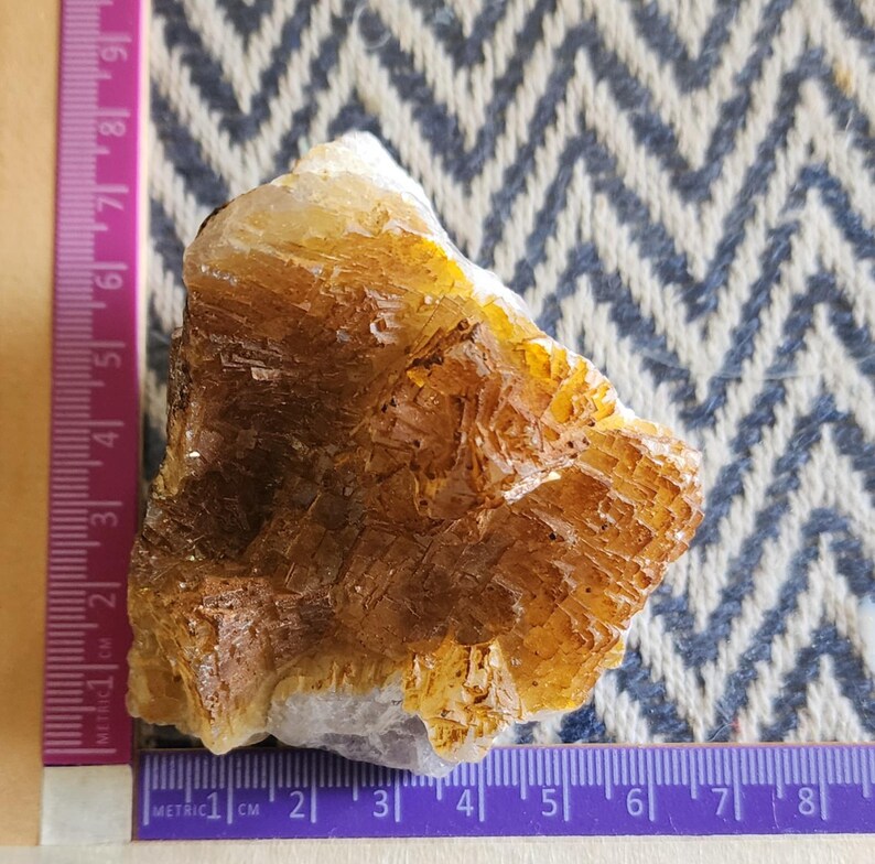 May include: A rough, natural citrine crystal with a layered, textured appearance. The crystal displays shades of golden yellow and brown, with a hint of white at the base. A purple and white metric ruler is placed next to the crystal, with measurements in centimeters.