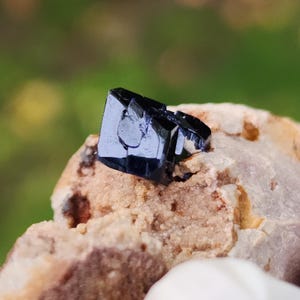 May include: A dark blue, crystalline mineral specimen with a reflective surface. The mineral is set on a rough, beige-colored rock, with a blurred green background. The mineral's geometric structure is clearly visible.