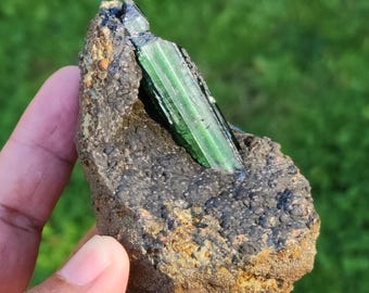 May include: A raw, natural green tourmaline crystal specimen embedded in a matrix of dark gray and brown rock. The tourmaline is a vibrant, translucent green, with visible striations. The rock has a rough, textured surface, with areas of dark gray and golden brown.
