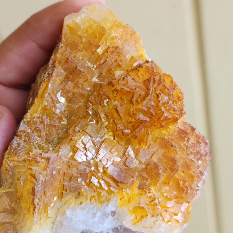 May include: A close-up of a rough, natural mineral specimen. The stone features a complex crystalline structure with a mix of golden yellow and orange hues. The base of the stone has a white and translucent appearance.