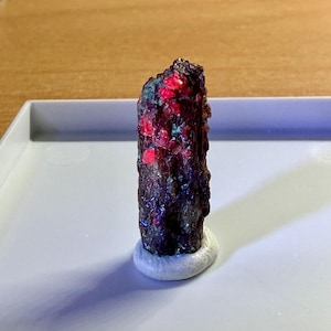 May include: A raw, elongated mineral specimen featuring a dark, textured, metallic-looking exterior with vibrant, bright red crystalline inclusions scattered across its surface, standing upright on a small, circular white base.