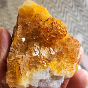 May include: A close-up of a rough, natural mineral specimen. The stone features a layered, crystalline structure with shades of yellow, orange, and brown. The base of the stone is a lighter, off-white color. The mineral is held in a hand.