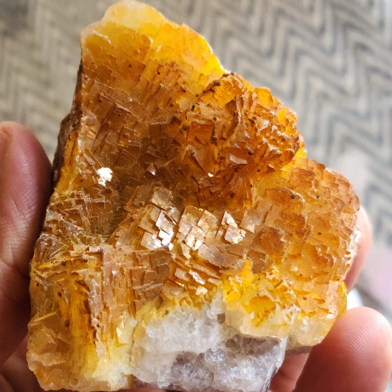 May include: A close-up of a rough, natural mineral specimen. The stone features a layered, crystalline structure with shades of yellow, orange, and brown. The base of the stone is a lighter, off-white color. The mineral is held in a hand.