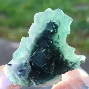 May include: A close-up of a fluorite crystal cluster. The crystal features a mix of translucent green and dark blue-green hues. The crystal's surface is composed of many small, geometric shapes.