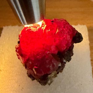 May include: A vibrant red mineral specimen illuminated by a light source. The crystalline structure of the mineral is visible, with a dark, textured base. The light source is a metallic object.