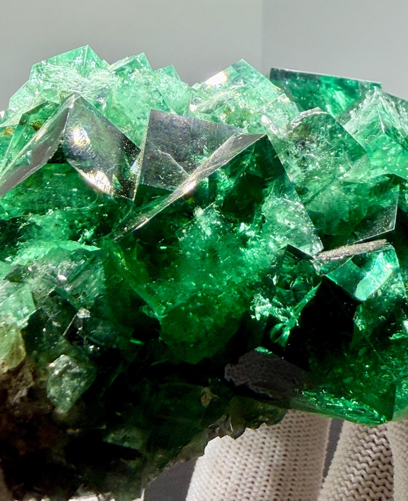 May include: A cluster of vibrant green fluorite crystals featuring sharp, geometric cubic structures with translucent surfaces that catch the light, displaying deep emerald hues and internal fractures throughout the mineral specimen.