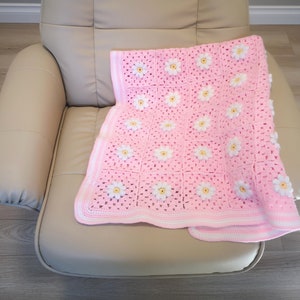 May include: A pink crocheted baby blanket with white daisy flowers. The blanket is folded and placed on a light brown leather armchair.